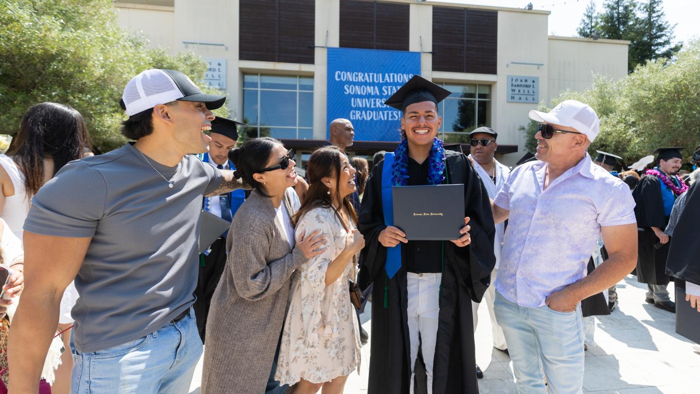 Student in graduation regalia holding a diploma with a group of four family members looking at them proud